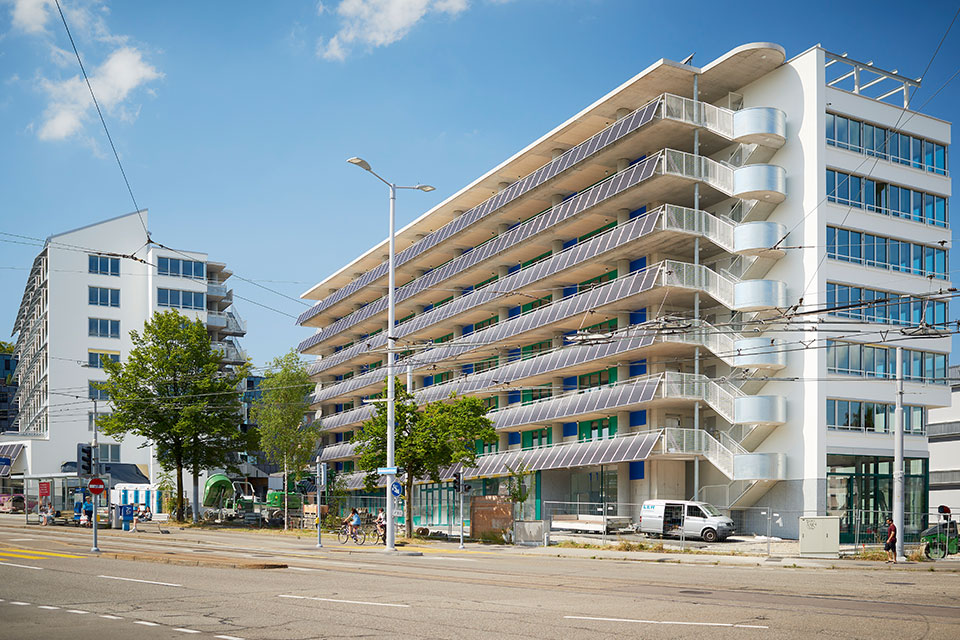 Vorderansicht des Guggach III Quartiers mit modernen Wohngebäuden, die mit Solarpanels ausgestattet sind, bei sonnigem Wetter.