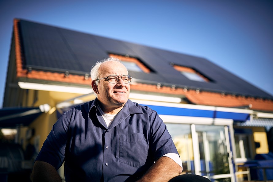 Ein Mann steht vor einem Haus und blickt in die Sonne. Auf dem Dach im Hintergrund sind Solarpanels zu sehen.